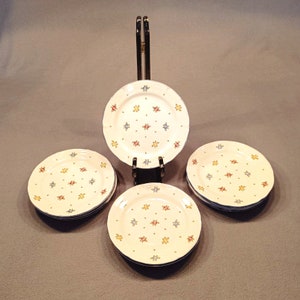 Pode incluir: Conjunto de quatro pratos de sobremesa de cerâmica branca com borda azul e um padrão de pequenas flores vermelhas, amarelas e azuis. Os pratos estão empilhados em um suporte preto.