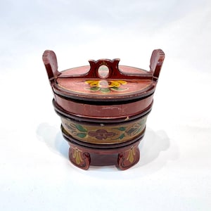 Op de afbeelding: Een klein, houten, rood, geel en groen geschilderd doosje met een deksel en een handvat. Het doosje heeft een bloemmotief en staat op drie pootjes.