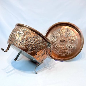 Può includere: Un piatto da portata in rame con coperchio, entrambi decorati con motivi di vite in rilievo. Il piatto ha tre gambe e una maniglia ad anello sul coperchio.