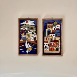 May include: Two framed ceramic wall plaques depicting colorful scenes of a Mediterranean village. The left plaque features a sailboat in a blue bay with a red sun and white clouds. The right plaque shows a multi-colored village with a yellow building and a red door.