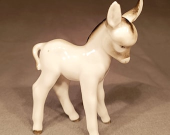 May include: A white porcelain figurine of a donkey foal. The foal is standing with its head down and its tail curled. The figurine has brown accents on its ears, mane, and hooves.