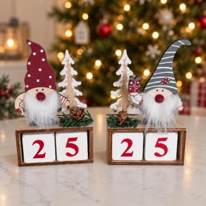 May include: Two wooden Christmas countdown block calendars featuring gnomes with white faux fur beards. One gnome wears a red polka-dot hat, the other a grey striped hat. Each sits on a wooden base with two white blocks displaying the number 25.