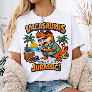 May include: White t-shirt with a cartoon orange Tyrannosaurus Rex wearing sunglasses and a Hawaiian shirt, holding a tropical drink. The shirt has the words "Vacasaurus" and "Time to get Jurassic!" in orange and blue.