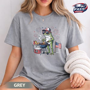 Könnte beinhalten: Graues T-Shirt mit einer Cartoon-Frosch, der eine Sonnenbrille und einen patriotischen Hut trägt und Burger auf einem Grill grillt. Eine amerikanische Flagge und Feuerwerk sind im Hintergrund. Das Shirt hat das Wort "GREY" unten.
