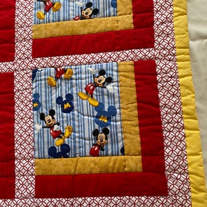 Pode incluir: Um edredom colorido com Mickey Mouse. O edredom tem um padr&atilde;o de listras azuis e brancas com ilustra&ccedil;&otilde;es do Mickey Mouse. Bordas vermelhas e douradas emolduram os quadrados do Mickey Mouse, com uma borda amarela ao redor da borda.