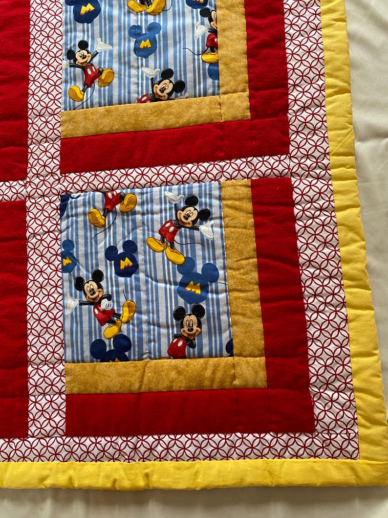 Pode incluir: Um edredom colorido com Mickey Mouse. O edredom tem um padr&atilde;o de listras azuis e brancas com ilustra&ccedil;&otilde;es do Mickey Mouse. Bordas vermelhas e douradas emolduram os quadrados do Mickey Mouse, com uma borda amarela ao redor da borda.