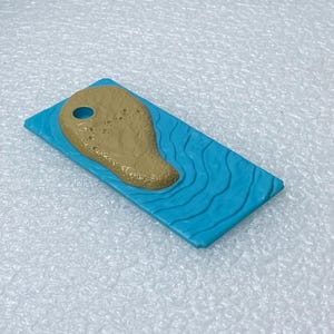 May include: A rectangular blue plastic piece with a molded wavy water texture. Attached to the surface is a tan, textured, island-shaped element featuring a single circular hole near the top edge, designed with a raised, grainy surface.