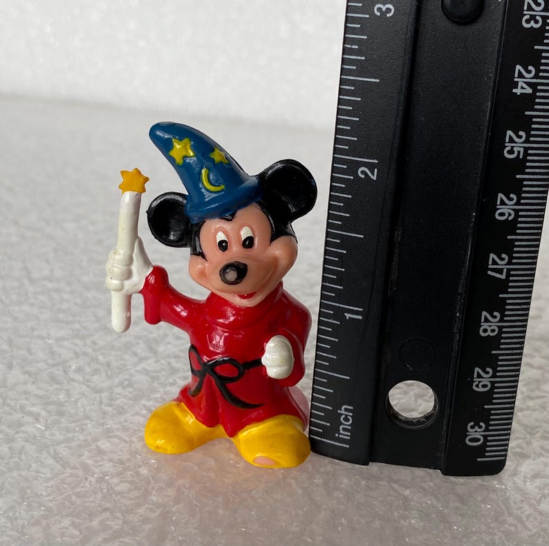 Pu&ograve; includere: Una piccola statuetta di Topolino vestito da stregone con una tunica rossa, cintura nera, scarpe gialle e un cappello blu con stelle gialle e una mezzaluna. Il personaggio tiene in mano una bacchetta bianca accanto a un righello nero.