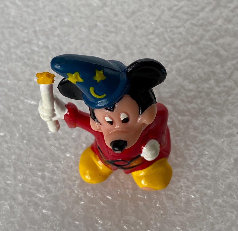 Pu&ograve; includere: Una piccola statuetta di un topo dei cartoni animati che indossa un cappello da mago blu con stelle e lune gialle, una tunica rossa e scarpe gialle. Il topo tiene una candela bianca in una mano e indossa guanti bianchi su entrambe le mani.