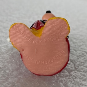 Pu&ograve; includere: Una piccola statuetta che mostra la pianta di due piedi di colore rosa chiaro. Il testo stampato recita WALT DISNEY PRODUCTIONS e MADE IN HONG KONG. La figura presenta accenti gialli sui bordi dei piedi e indumenti rossi visibili.