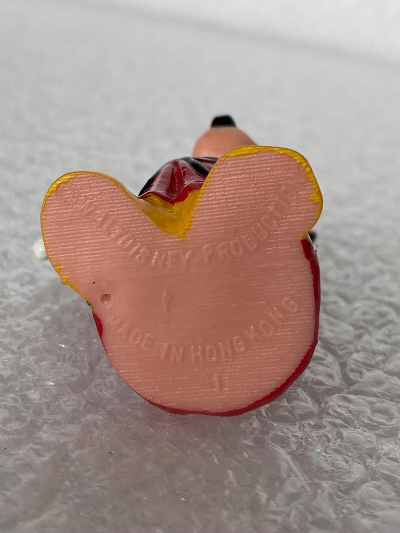Pu&ograve; includere: Una piccola statuetta che mostra la pianta di due piedi di colore rosa chiaro. Il testo stampato recita WALT DISNEY PRODUCTIONS e MADE IN HONG KONG. La figura presenta accenti gialli sui bordi dei piedi e indumenti rossi visibili.