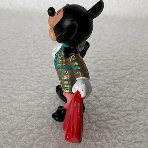 May include: A small figurine of a cartoon mouse character dressed in a gold and teal matador-style jacket with a white ruffled collar, white gloves, and black shoes, holding a bright red cape in one hand while looking upward in profile.