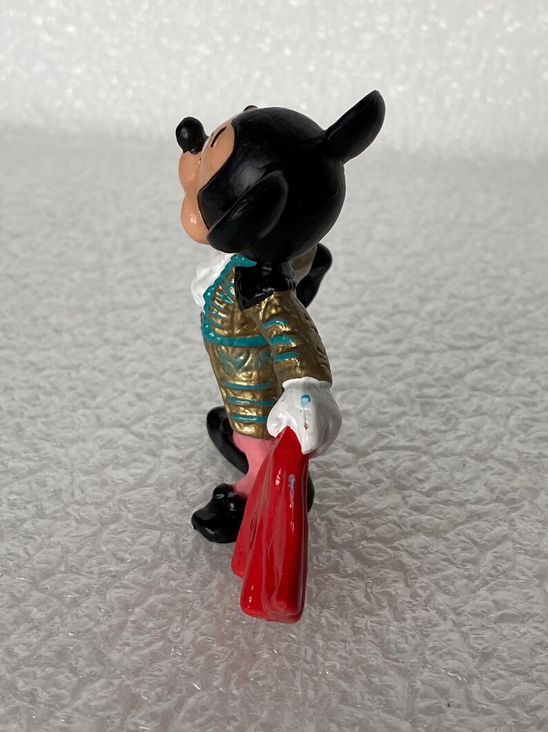 May include: A small figurine of a cartoon mouse character dressed in a gold and teal matador-style jacket with a white ruffled collar, white gloves, and black shoes, holding a bright red cape in one hand while looking upward in profile.
