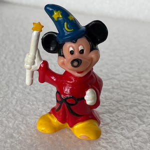 Pu&ograve; includere: Una piccola statuetta di un topo che indossa un cappello da mago blu con stelle gialle e una luna crescente, una tunica rossa con una cintura nera e scarpe gialle. Il personaggio tiene in mano una bacchetta bianca con una stella gialla.