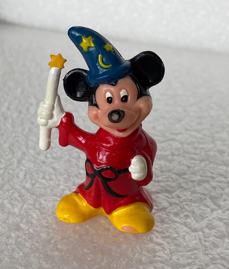Pu&ograve; includere: Una piccola statuetta di un topo che indossa un cappello da mago blu con stelle gialle e una luna crescente, una tunica rossa con una cintura nera e scarpe gialle. Il personaggio tiene in mano una bacchetta bianca con una stella gialla.