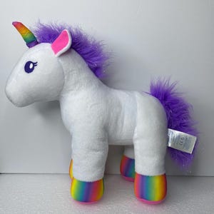 May include: A white plush unicorn toy featuring a rainbow-colored horn, bright purple mane and tail, and rainbow-striped hooves. The unicorn has large dark eyes with long eyelashes and pink inner ears, with a white product tag attached to the rear.