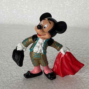 May include: A small figurine of Mickey Mouse dressed in a gold and teal matador outfit with a white ruffled shirt. The character holds a black hat in one hand and a bright red cape in the other, wearing pink leggings and black shoes.
