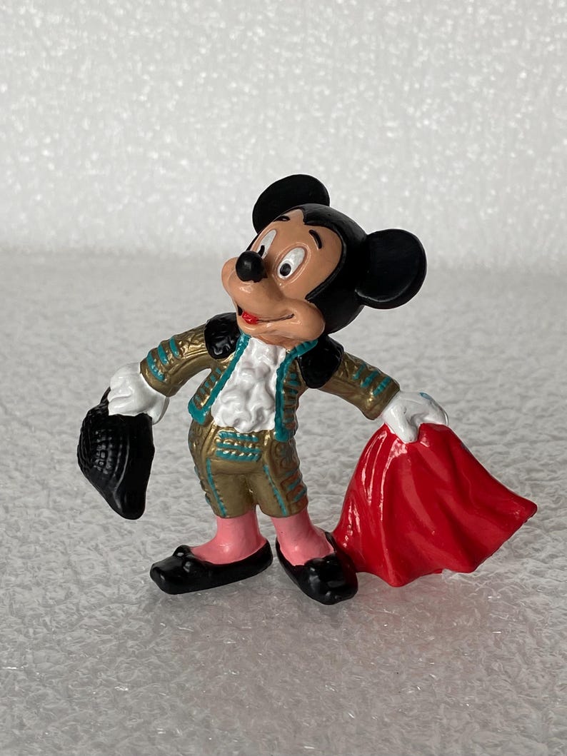 May include: A small figurine of Mickey Mouse dressed in a gold and teal matador outfit with a white ruffled shirt. The character holds a black hat in one hand and a bright red cape in the other, wearing pink leggings and black shoes.