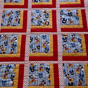 Pode incluir: Um edredom feito &agrave; m&atilde;o com um padr&atilde;o repetido do Mickey Mouse em um fundo listrado azul e branco. Os quadrados s&atilde;o emoldurados com tecido dourado, vermelho e branco estampado. O edredom &eacute; um design de patchwork.
