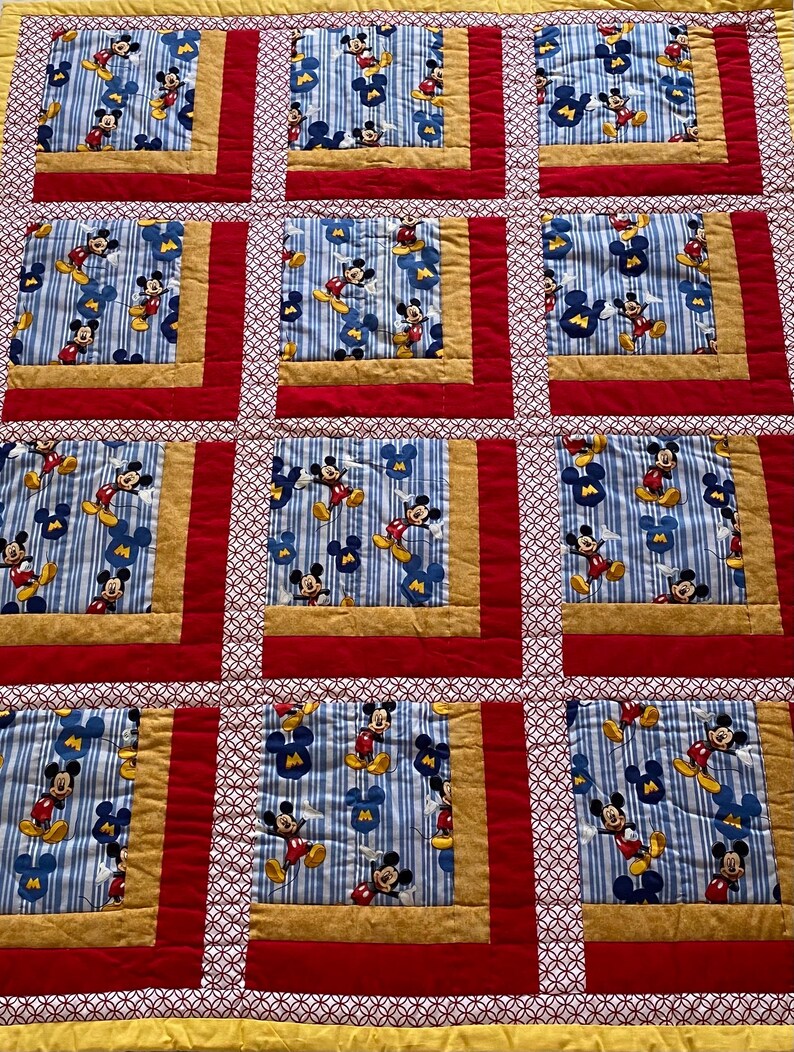 Pode incluir: Um edredom feito &agrave; m&atilde;o com um padr&atilde;o repetido do Mickey Mouse em um fundo listrado azul e branco. Os quadrados s&atilde;o emoldurados com tecido dourado, vermelho e branco estampado. O edredom &eacute; um design de patchwork.