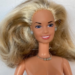 May include: A doll with long blonde hair, blue eyes, and a smiling expression. The doll wears small grey stud earrings and a thin silver-toned necklace featuring three small metallic beads centered at the front of the neck against a tan skin tone.