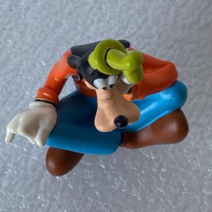 以下が含まれることがあります： ディズニーの漫画のキャラクター、グーフィーのフィギュア。オレンジ色のシャツ、青いズボン、茶色の靴、黒い帽子、緑の帽子を着用。白い手袋が見え、顔には大きな笑顔が浮かんでいます。