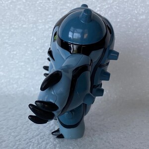 May include: A blue and black action figure of a fictional character. The figure has a rounded body with black stripes and accents. It has multiple black protrusions and green eyes. The figure is standing on a white surface.