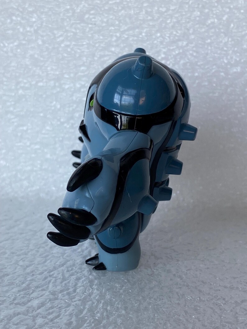 May include: A blue and black action figure of a fictional character. The figure has a rounded body with black stripes and accents. It has multiple black protrusions and green eyes. The figure is standing on a white surface.