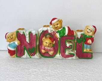 May include: A ceramic candle holder shaped like the word NOEL in red letters with white snow accents. Four teddy bears wearing winter clothing and Santa hats are positioned around the letters. Two gold candle cups sit on top.
