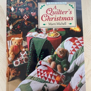 May include: A book titled "A Quilter's Christmas" by Marti Michell, a Rodale Quilt Book. The cover features a cozy living room scene with a decorated Christmas tree, a fireplace, and two teddy bears sitting on a quilt.