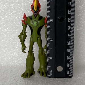 May include: A green and black action figure with red accents on the head and shoulders, featuring a central chest emblem. The figure stands next to a black ruler, showing a height of approximately 4.25 inches from the base to the top of the head.