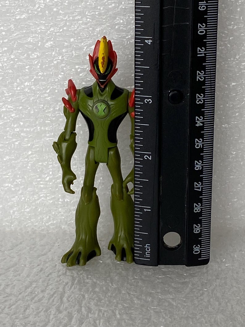 May include: A green and black action figure with red accents on the head and shoulders, featuring a central chest emblem. The figure stands next to a black ruler, showing a height of approximately 4.25 inches from the base to the top of the head.