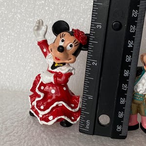 May include: A Minnie Mouse figurine wearing a red dress with white polka dots and a red flower on her head, positioned next to a black ruler showing a height of approximately 3 inches for the character.