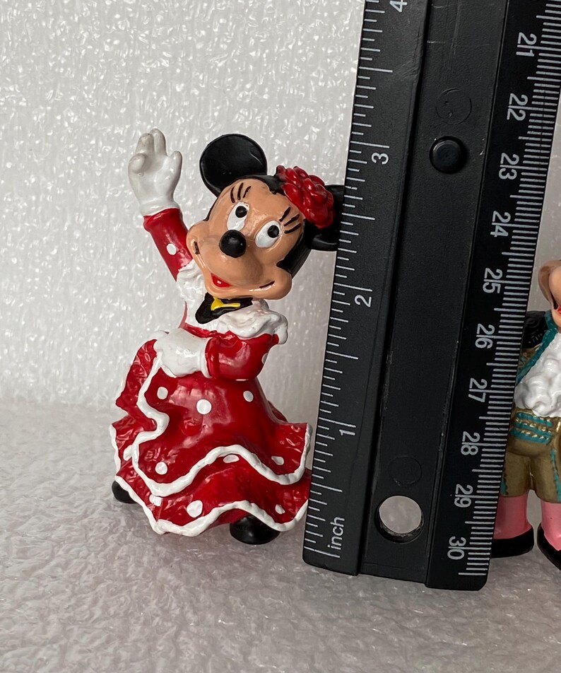 May include: A Minnie Mouse figurine wearing a red dress with white polka dots and a red flower on her head, positioned next to a black ruler showing a height of approximately 3 inches for the character.