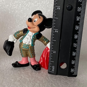 May include: A Mickey Mouse figurine dressed in a gold and teal matador outfit with a red cape, standing next to a black ruler that shows the figure is approximately 3 inches tall. The character has black ears, a peach face, and black shoes.