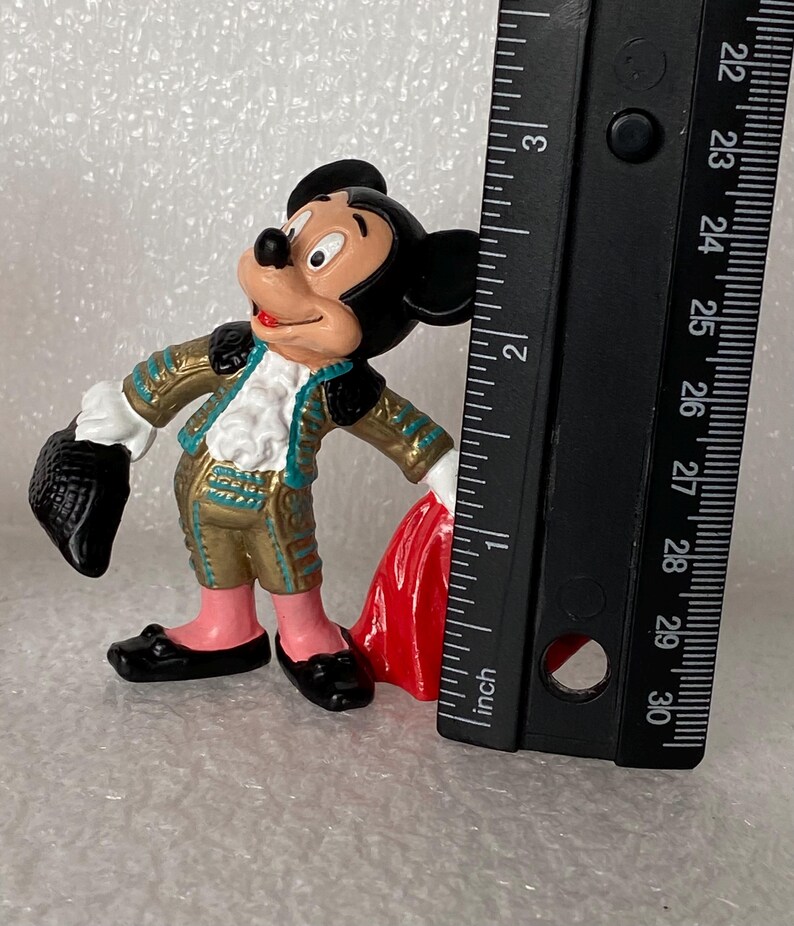 May include: A Mickey Mouse figurine dressed in a gold and teal matador outfit with a red cape, standing next to a black ruler that shows the figure is approximately 3 inches tall. The character has black ears, a peach face, and black shoes.