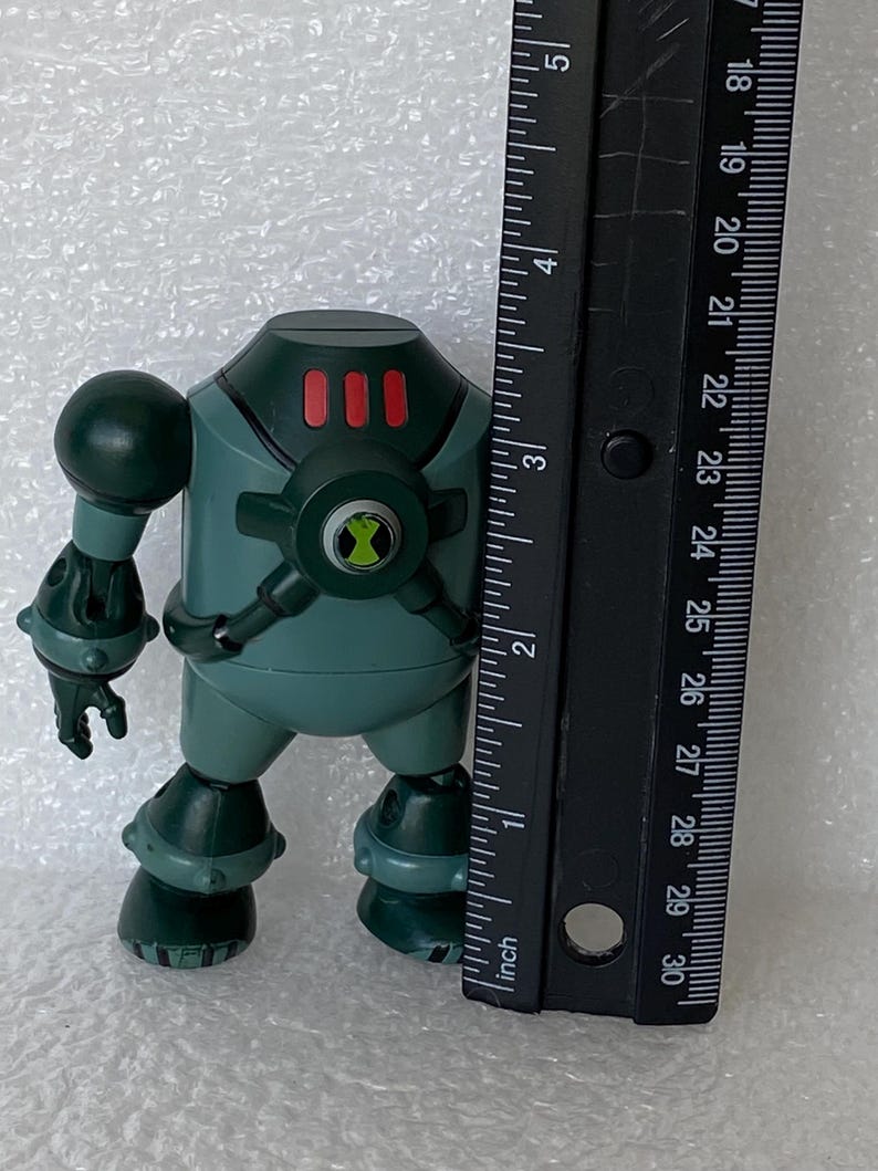 May include: A teal and dark green action figure of a robot-like character, approximately 10 cm tall. The figure has a round body, a head with three red rectangles, and a circular emblem. A black ruler is placed next to the figure.