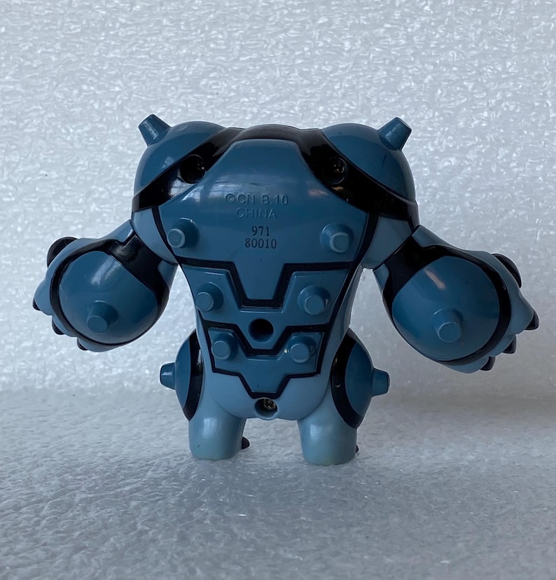 May include: A blue and black plastic toy figure with a stylized, robotic design. The figure has a rounded body with protruding details and black accents. The text "OCII B-10 CHINA 971 80010" is printed on the back.