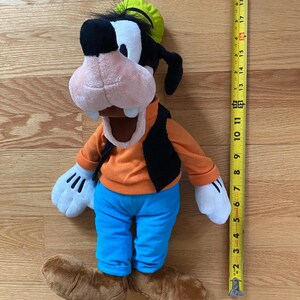 Może przedstawiać: Pluszowa zabawka przedstawiająca postać Goofy'ego w limonkowym kapeluszu, pomarańczowej koszulce z długim rękawem, czarnej kamizelce, niebieskich spodniach i brązowych butach. Zabawka stoi obok ż&oacute;łtej miarki wskazującej 51 centymetr&oacute;w wysokości.