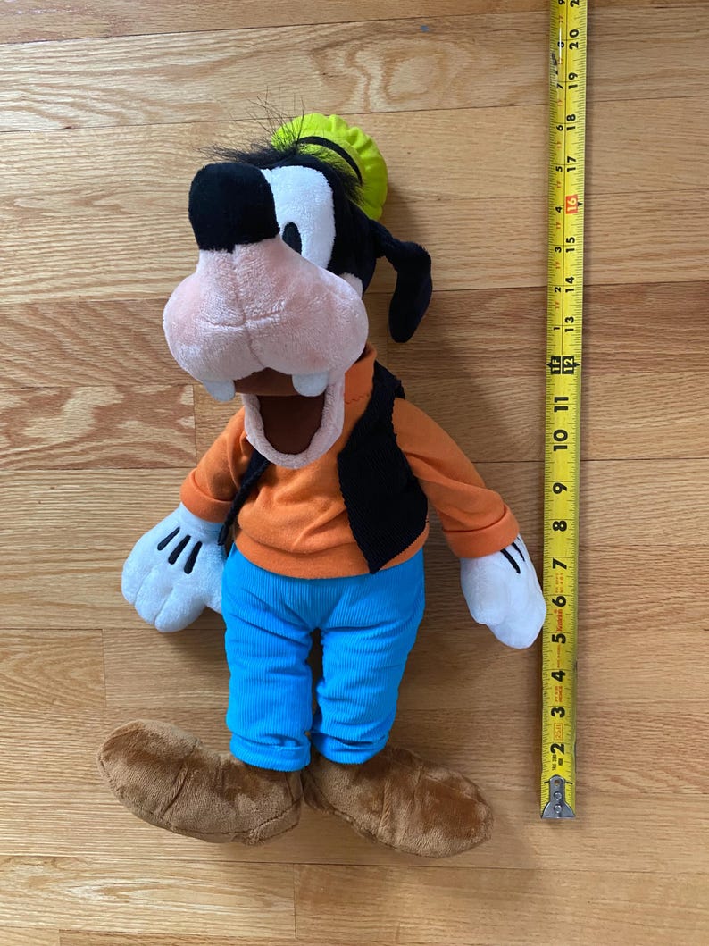 Może przedstawiać: Pluszowa zabawka przedstawiająca postać Goofy'ego w limonkowym kapeluszu, pomarańczowej koszulce z długim rękawem, czarnej kamizelce, niebieskich spodniach i brązowych butach. Zabawka stoi obok ż&oacute;łtej miarki wskazującej 51 centymetr&oacute;w wysokości.