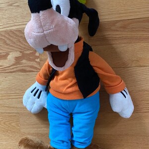 Może przedstawiać: Pluszowa zabawka przedstawiająca postać Goofy'ego z czarno-białą głową, zielonym kapeluszem, pomarańczową koszulką z długim rękawem, czarną kamizelką, jaskrawoniebieskimi spodniami, białymi rękawiczkami i brązowymi butami. Postać ma otwarte usta.