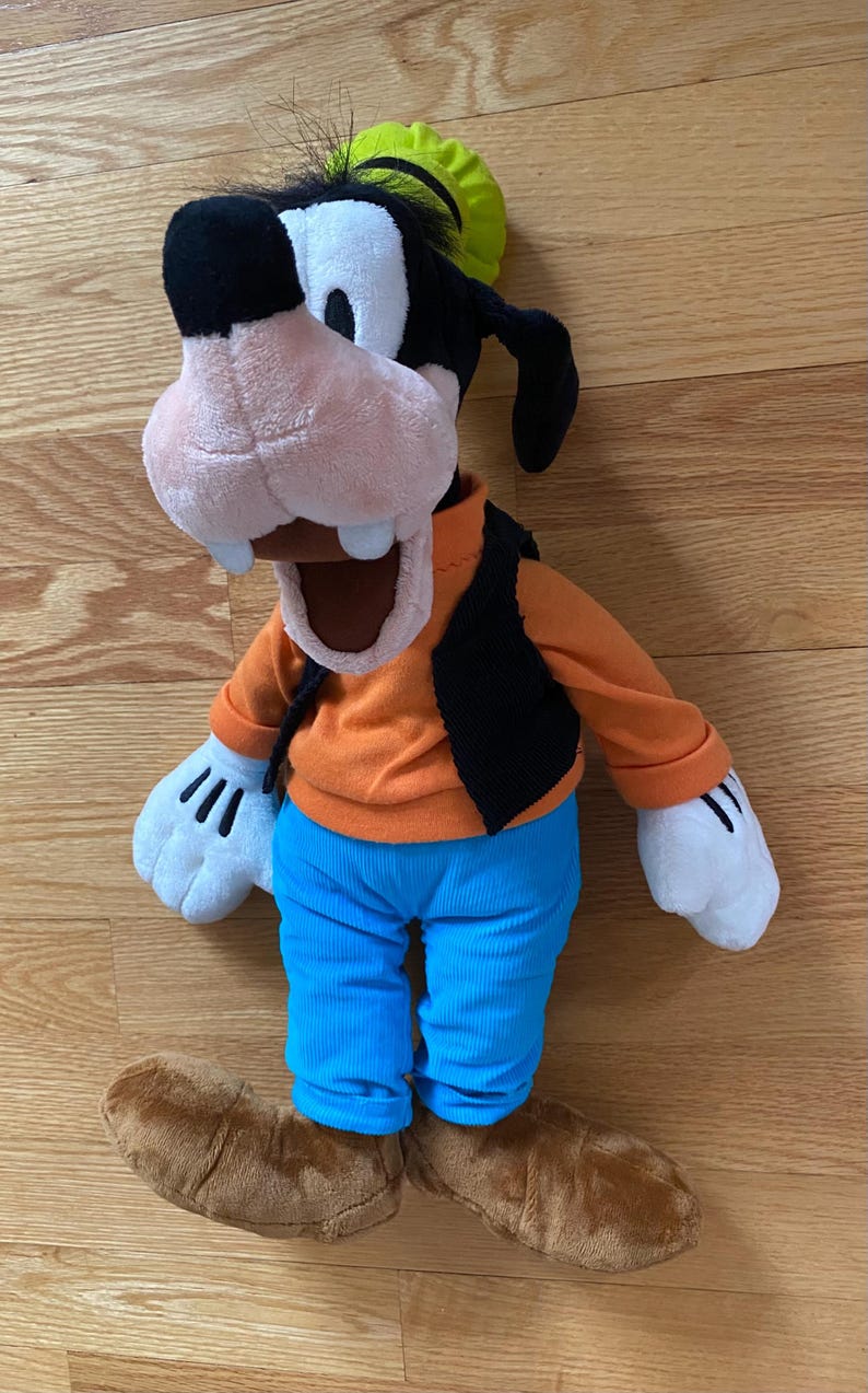 Może przedstawiać: Pluszowa zabawka przedstawiająca postać Goofy'ego z czarno-białą głową, zielonym kapeluszem, pomarańczową koszulką z długim rękawem, czarną kamizelką, jaskrawoniebieskimi spodniami, białymi rękawiczkami i brązowymi butami. Postać ma otwarte usta.