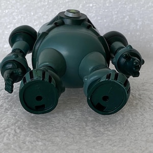 May include: A teal toy robot with a rounded body, cylindrical arms, and circular feet. The robot has a small, circular emblem on its head. The toy is made of plastic and has a matte finish.