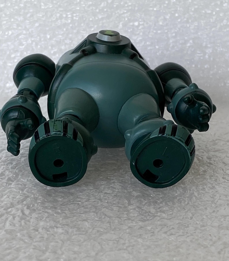 May include: A teal toy robot with a rounded body, cylindrical arms, and circular feet. The robot has a small, circular emblem on its head. The toy is made of plastic and has a matte finish.