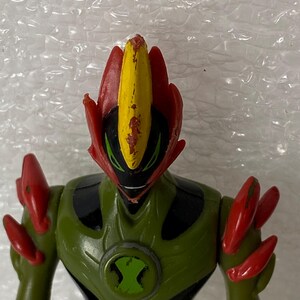May include: A plastic action figure of a character with a green and black body, red shoulder spikes, and a head featuring a central yellow crest with red accents. The chest displays a circular green and black emblem with a stylized hourglass shape.