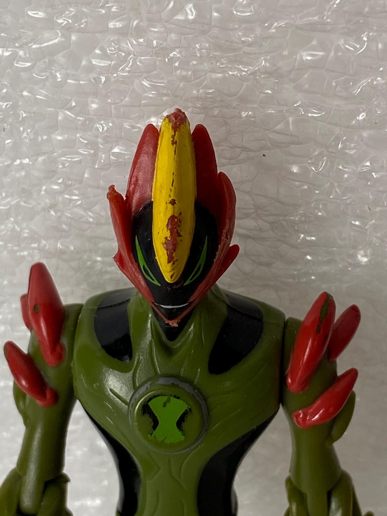 May include: A plastic action figure of a character with a green and black body, red shoulder spikes, and a head featuring a central yellow crest with red accents. The chest displays a circular green and black emblem with a stylized hourglass shape.