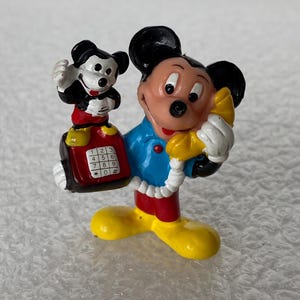 May include: A small, painted figurine of Mickey Mouse wearing a blue shirt, red shorts, and yellow shoes. Mickey holds a yellow telephone receiver to their ear and a red push-button telephone base with a smaller Mickey figure standing on top.