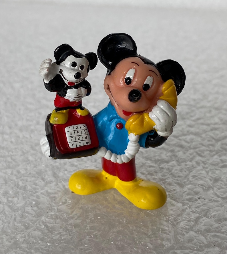 May include: A small, painted figurine of Mickey Mouse wearing a blue shirt, red shorts, and yellow shoes. Mickey holds a yellow telephone receiver to their ear and a red push-button telephone base with a smaller Mickey figure standing on top.