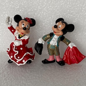May include: Two painted figurines of cartoon mice. One mouse wears a red dress with white polka dots and a red hair accessory. The other mouse wears a gold and teal matador outfit, holding a black hat in one hand and a red cape in the other.