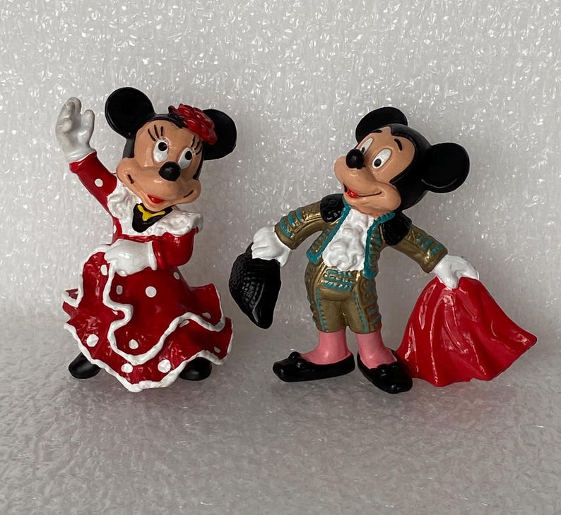 May include: Two painted figurines of cartoon mice. One mouse wears a red dress with white polka dots and a red hair accessory. The other mouse wears a gold and teal matador outfit, holding a black hat in one hand and a red cape in the other.